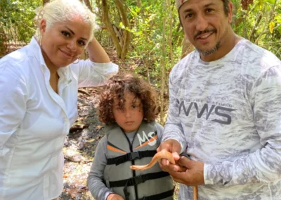 A man holds a snake as a child in a life jacket curiously watches, surrounded by lush greenery in a tropical setting