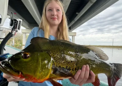 A person holds a large, vibrant green peacock bass while sitting in a boat under a concrete structure with water and cloudy skies in the background