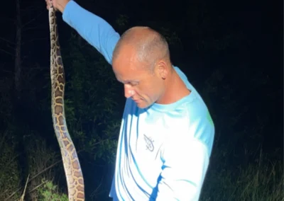A person holds a long, patterned snake in a dark, outdoor setting surrounded by grass and trees