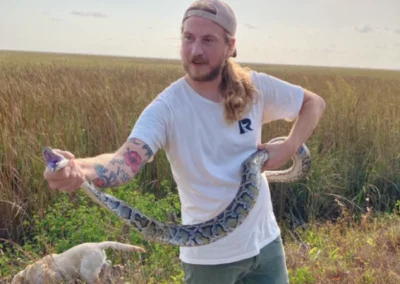 A person with long hair holds a large snake in an open field, with a dog visible in the background. The sky is partly cloudy
